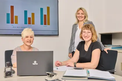 Drei nette Mitarbeiterinnen sitzen am Tisch mit Laptop und Büromaterial und lächeln freundlich in die Kamera.